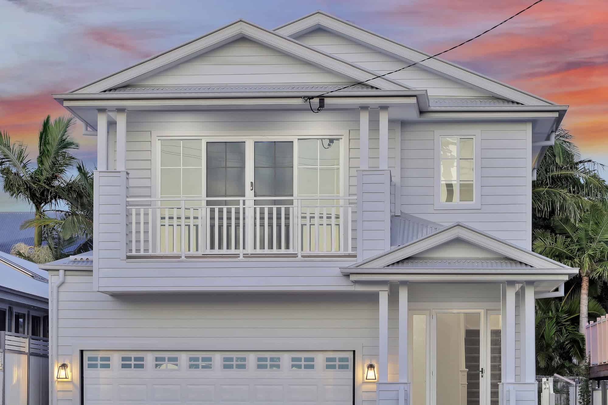 Two-storey hamptons-style Brisbane knockdown rebuild with weatherboard facade, upstairs balcony and double garage at dusk